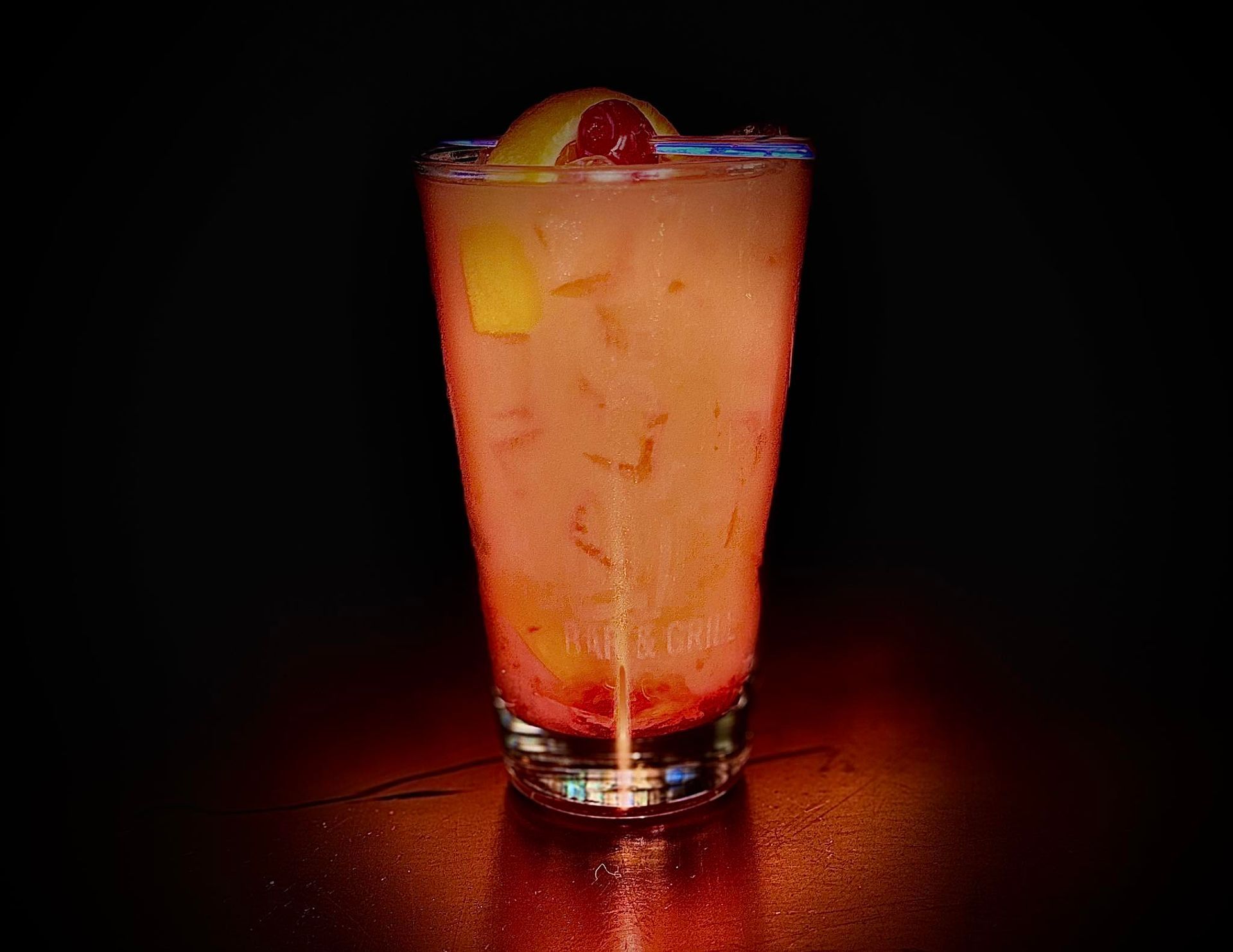 Tall glass of pink cocktail with ice, lemon slice, and cherry garnish, on a dark red surface.