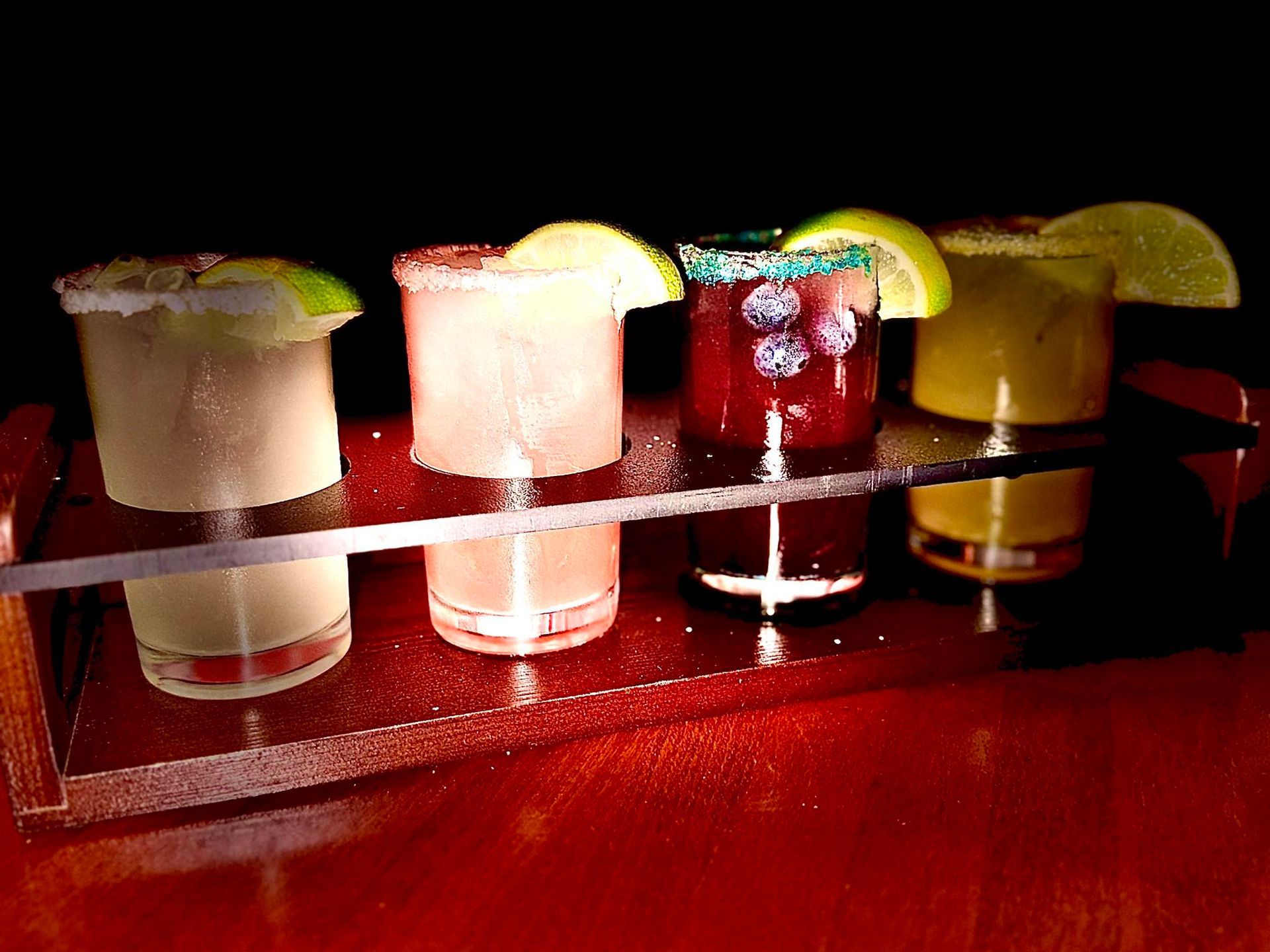 Four colorful cocktails in shot glasses on a tray, each with a lime wedge garnish.