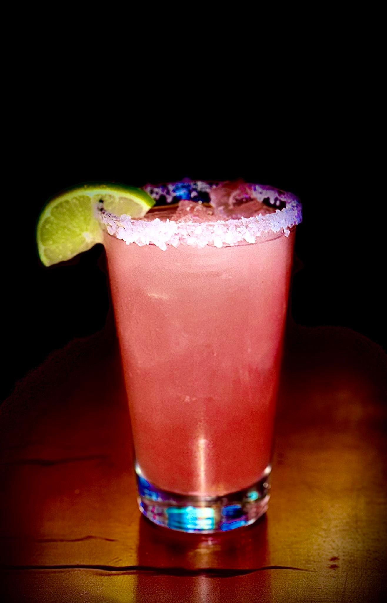Pink cocktail in a highball glass, salted rim, lime wedge garnish, dark background.