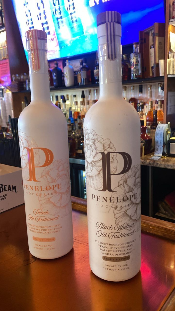 Two Penelope bourbon bottles on a bar, one with orange label, one with black label.