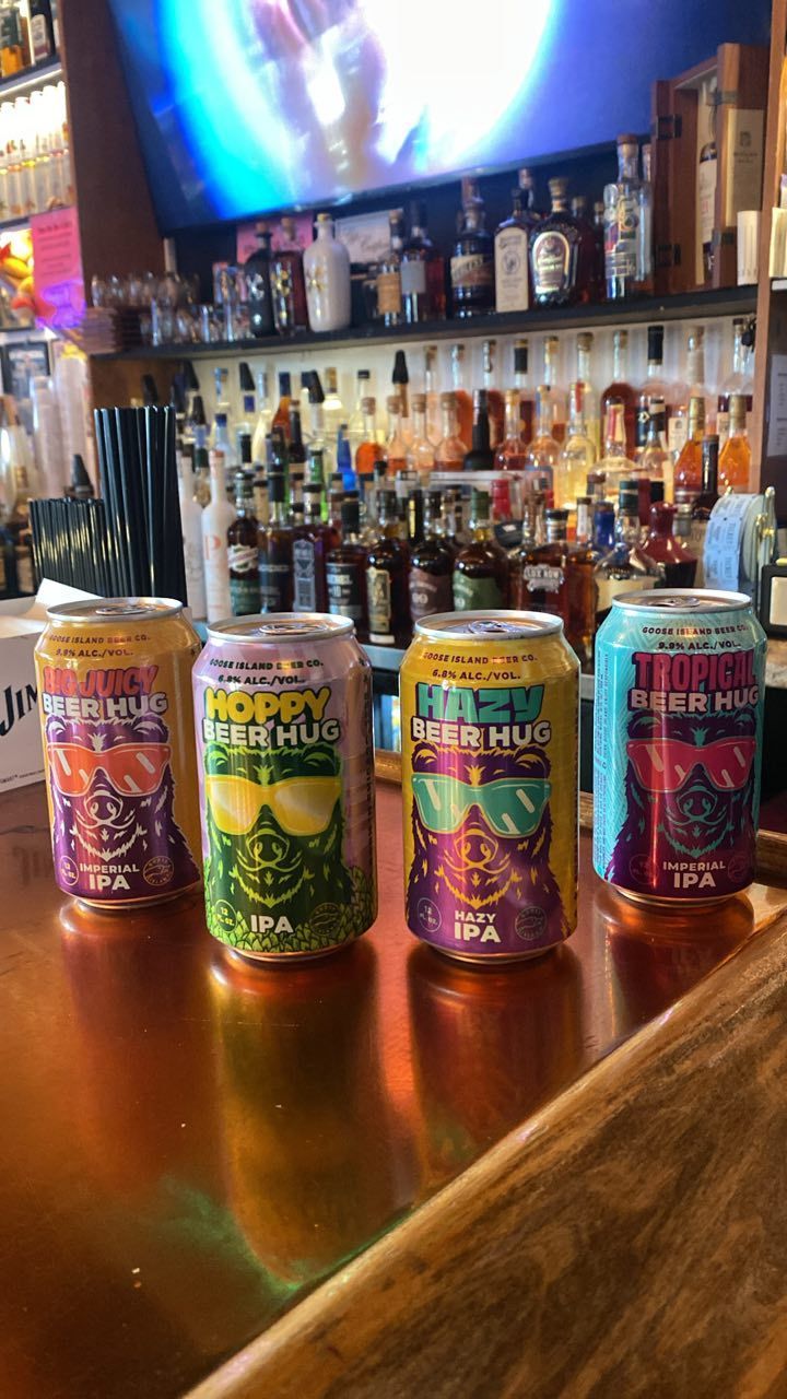 Four beer cans lined up on a bar with a blurred background of liquor bottles and a bar mirror.
