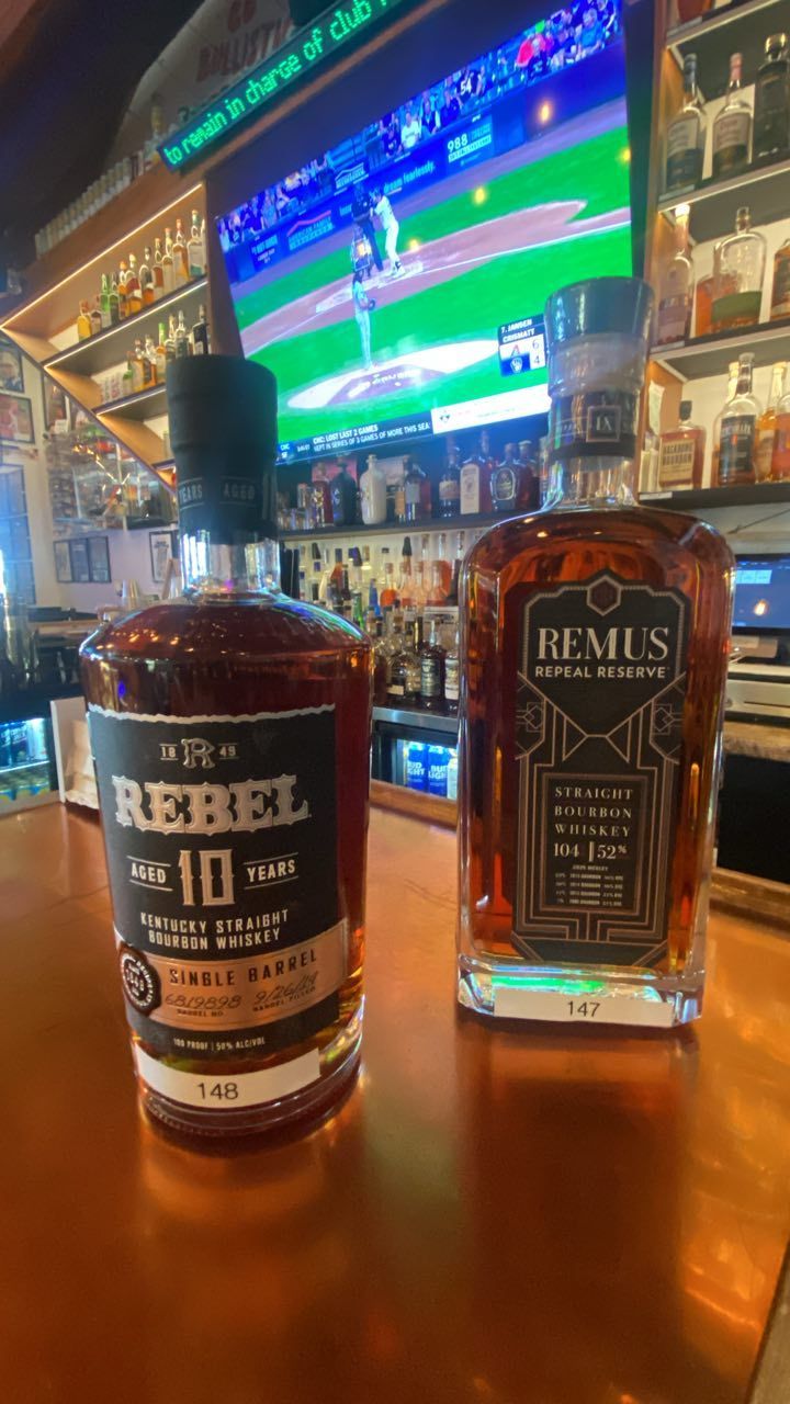 Two bottles of whiskey on a bar: Rebel 10 year and Remus Bourbon, with a TV in the background.