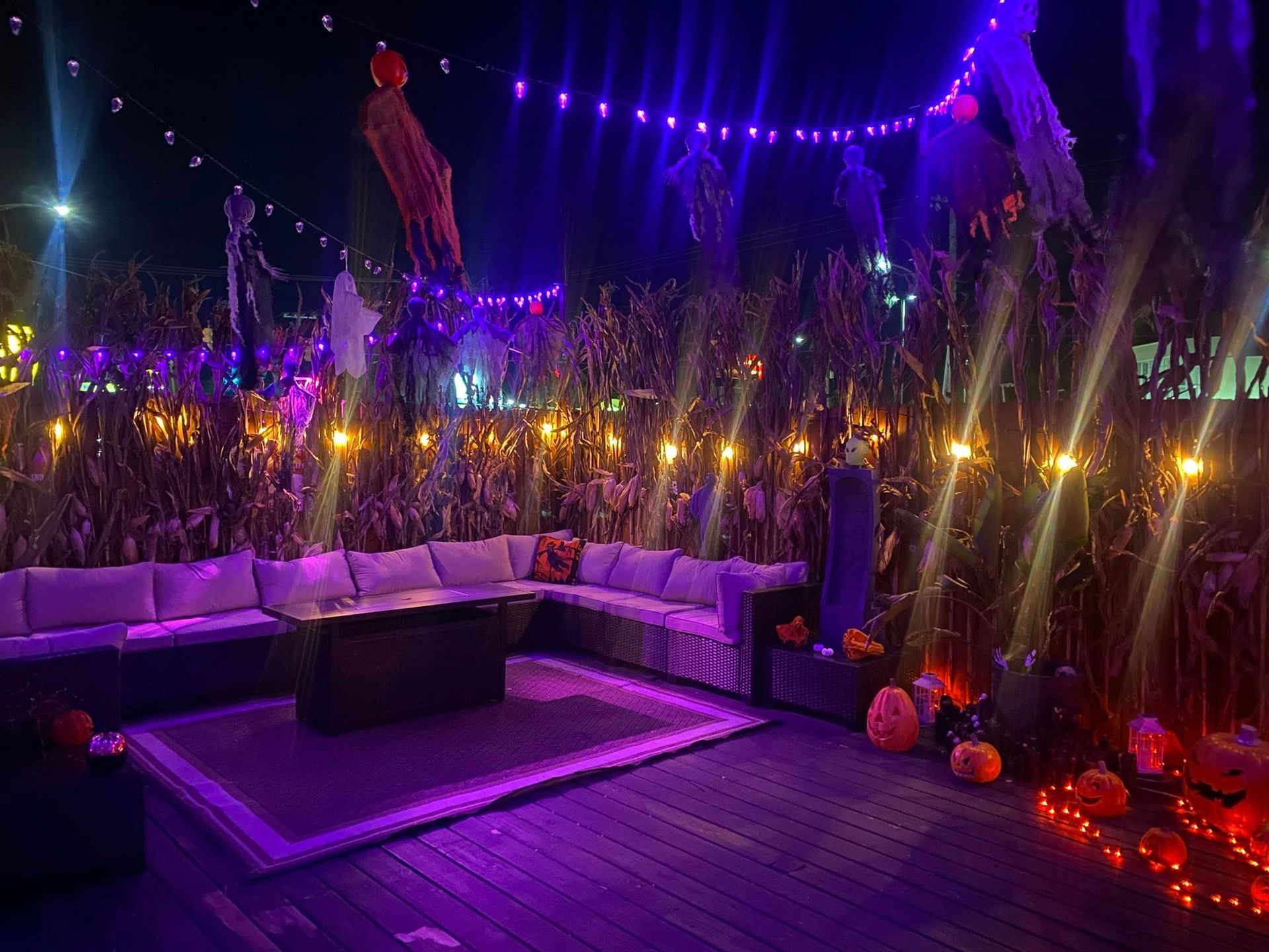 Halloween-themed outdoor lounge with purple lighting, pumpkins, and a fire pit, surrounded by corn stalks.