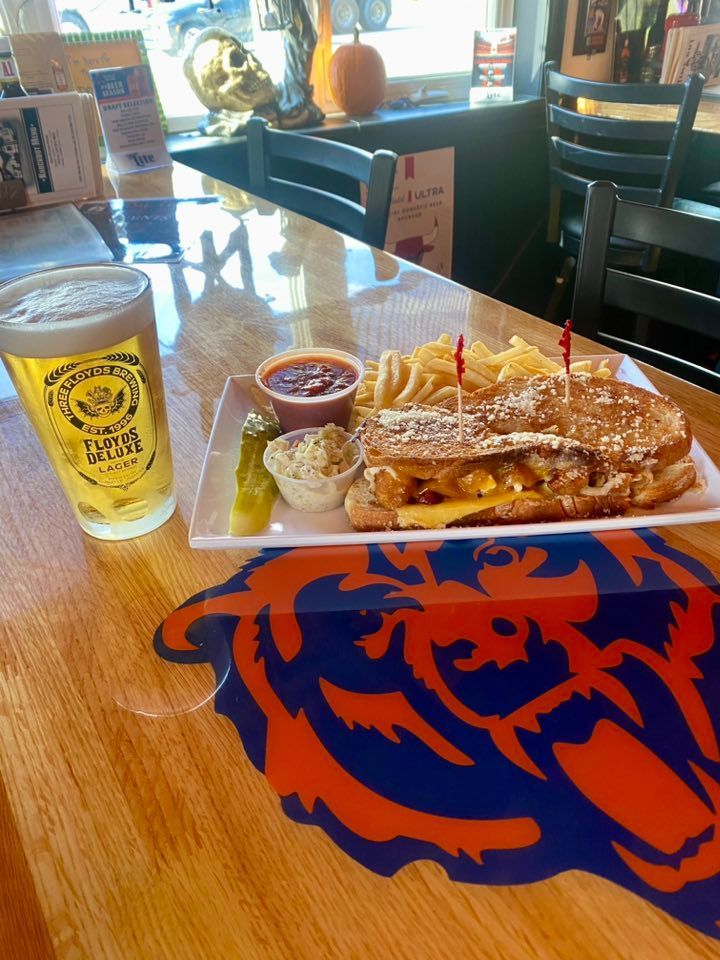 Grilled sandwich with fries, coleslaw, pickle, and beer on a table with a sports logo.