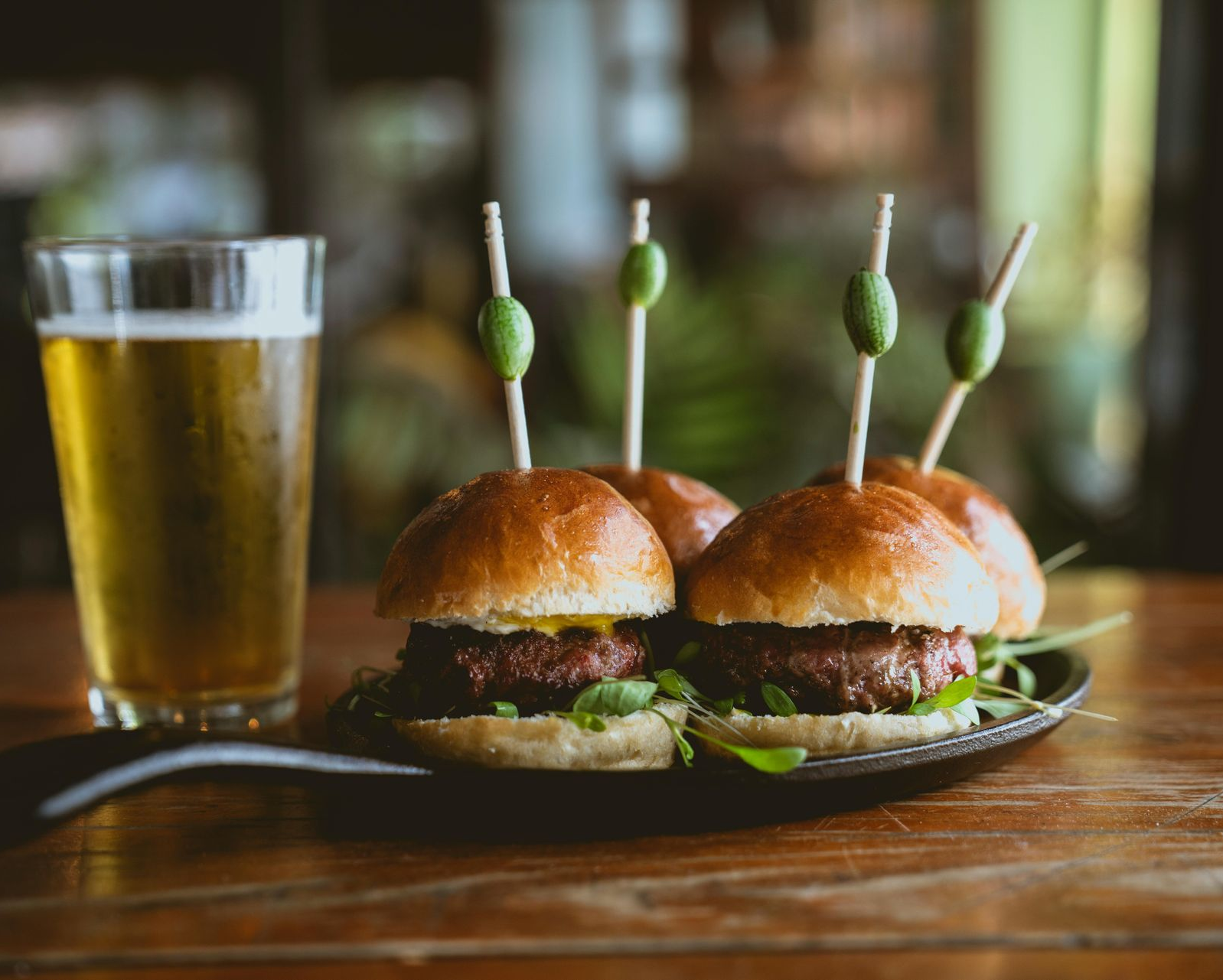 Mini burgers on plate, with olive-topped skewers, beside a glass of beer on a wooden table.