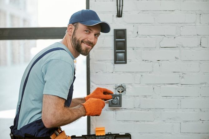 Electrician in blue shirt and cap tightening a wall outlet with orange gloves in a bright garage.