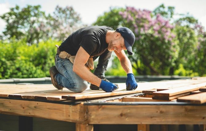 Worker kneeling on a rooftop, fastening wooden boards with a hammer, outdoors in sunlight