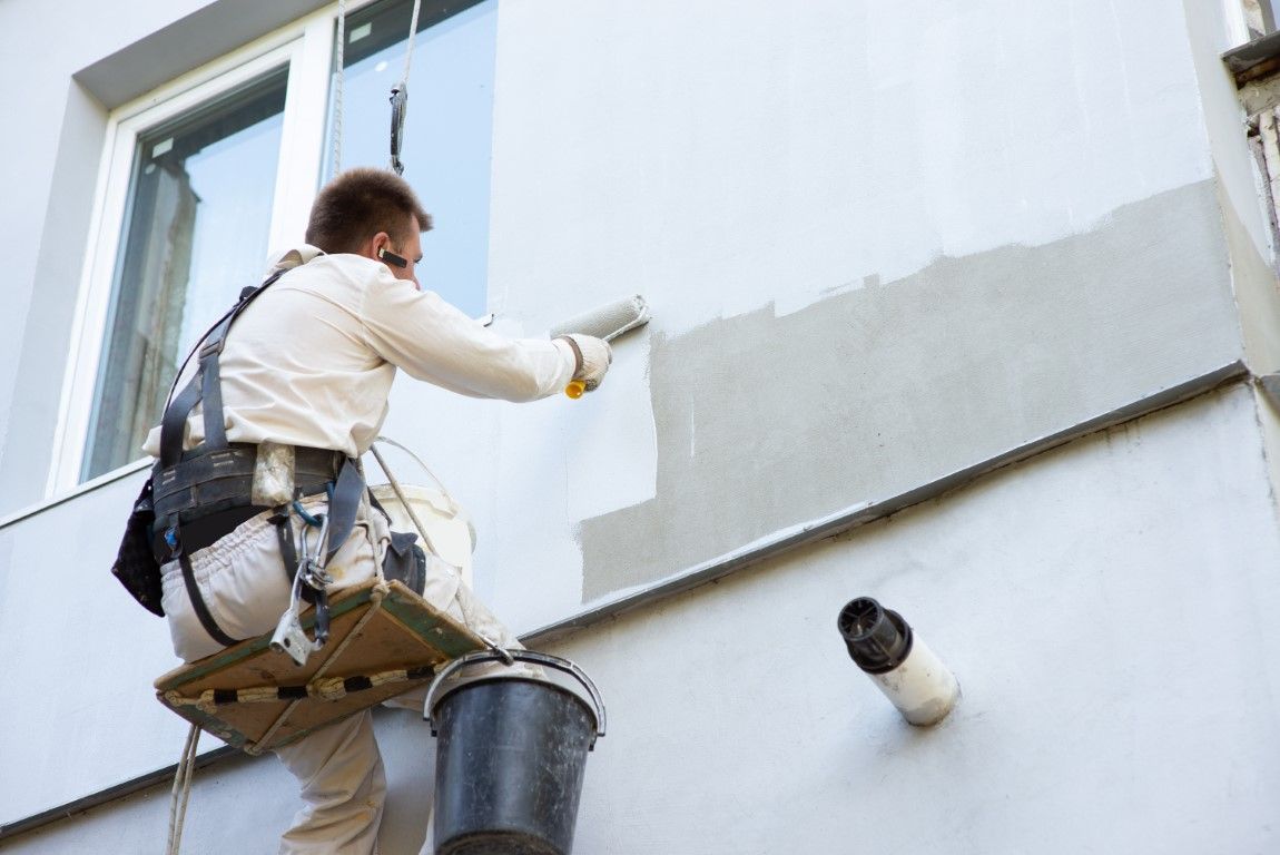 Painter on a scaffold painting a white exterior wall with gray primer near a window