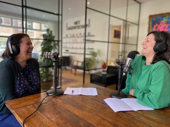 Shoshanna en Galit lachen tijdens een podcastopname. Ze zitten aan een tafel, met koptelefoons op en microfoons voor zich.