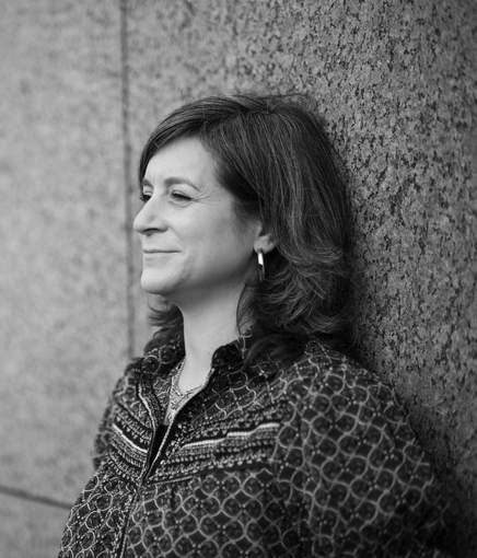 Galit leunt tegen een muur en kijkt vooruit met een lichte glimlach. Ze draagt een blouse met een patroon.