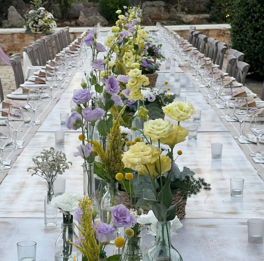 Lungo tavolo da esterno apparecchiato per un pasto, decorato con fiori in vasi di vetro.