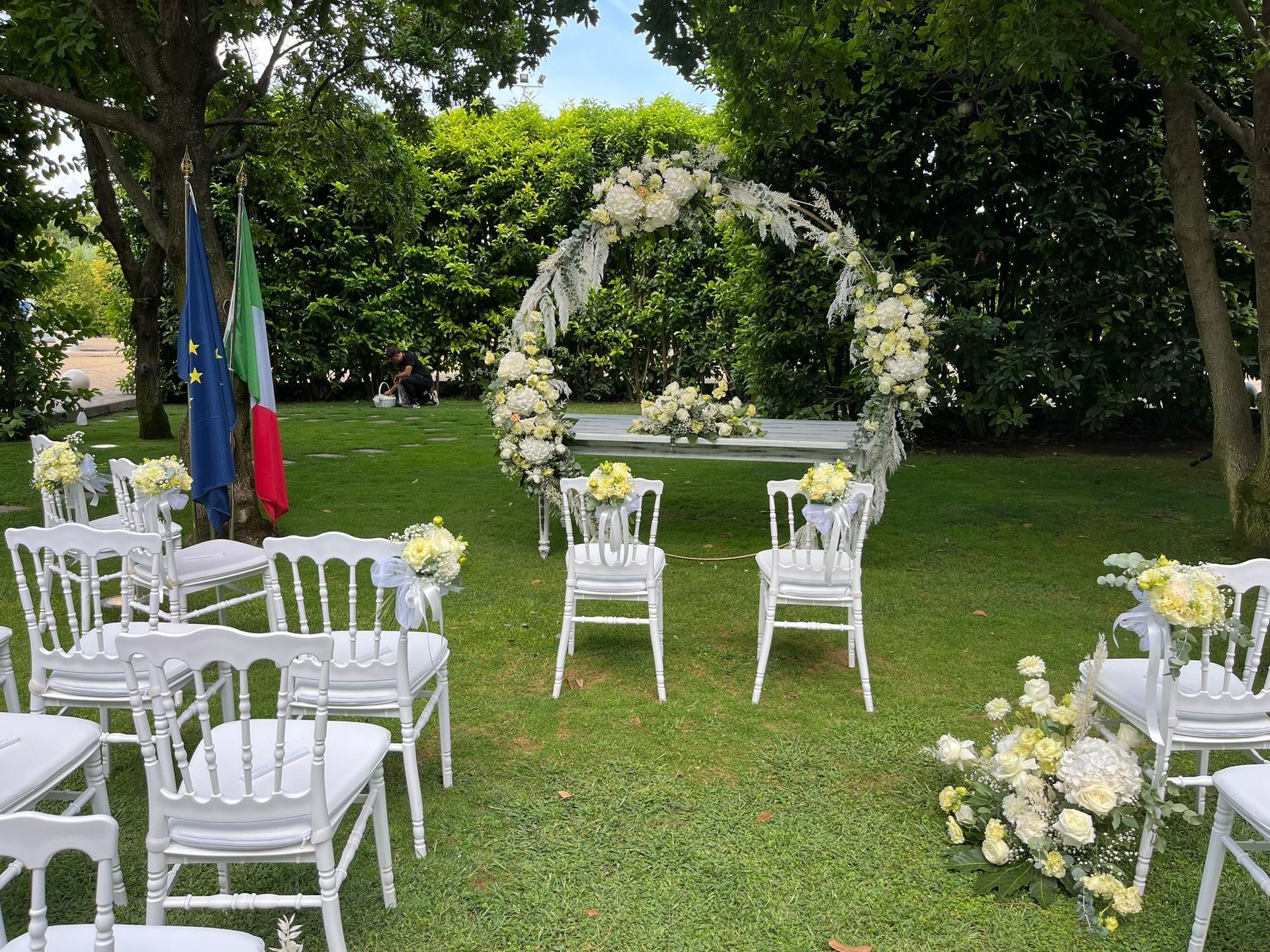 Allestimento per matrimonio all'aperto con sedie bianche, arco floreale e bandiere su un prato erboso.