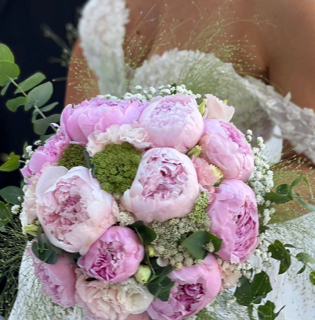 Primo piano di un bouquet di peonie rosa tenuto in mano da una persona che indossa un abito bianco.