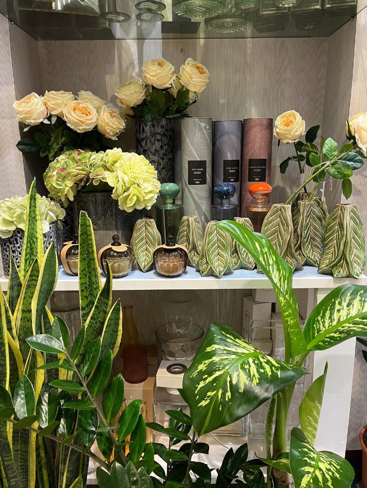 Esposizione di fiori e piante in vaso su scaffali. Include rose, ortensie, piante serpente e vasi decorati.