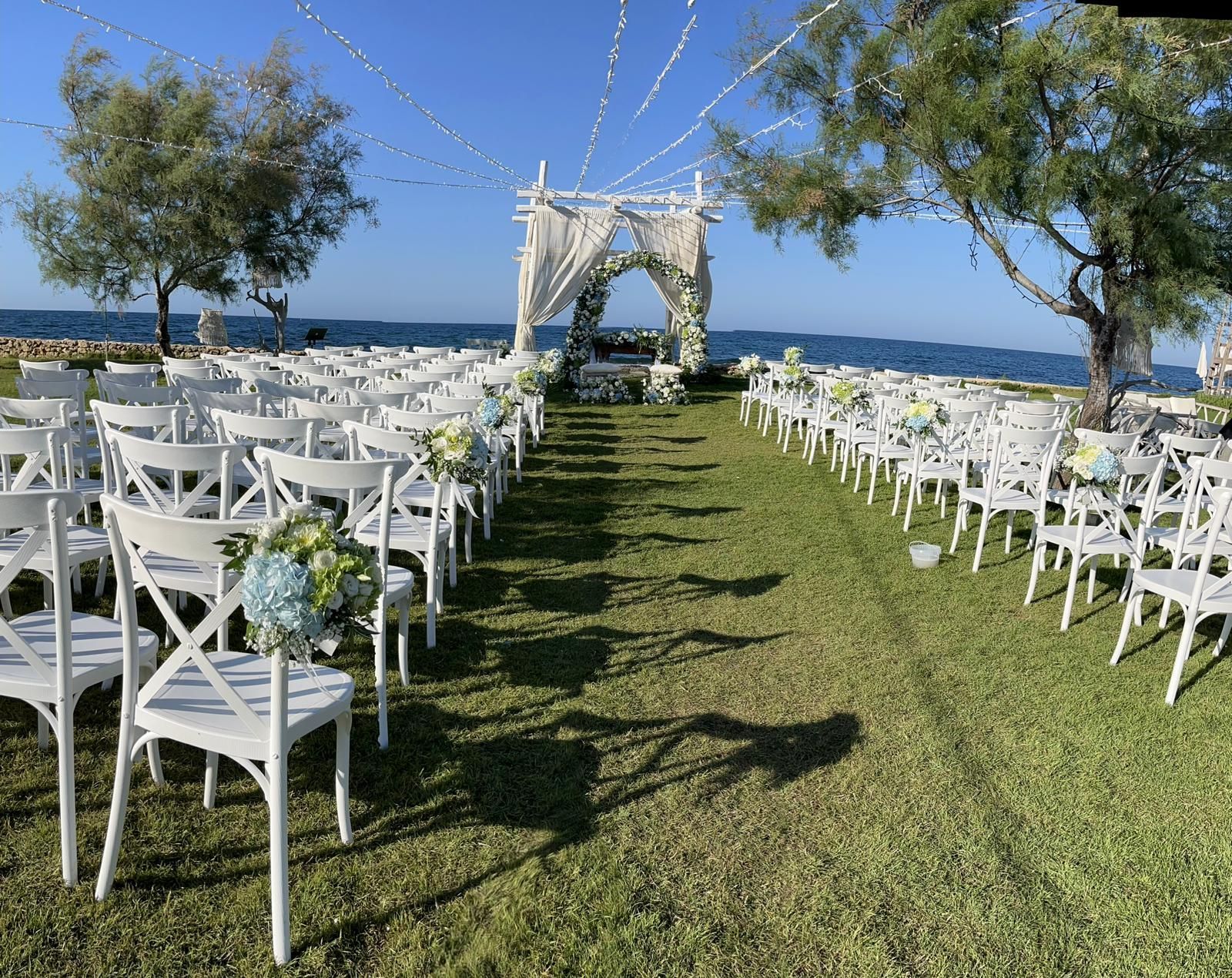 Cerimonia nuziale allestita su prato verde con vista sull'oceano. Sedie bianche, arco e decorazioni floreali.