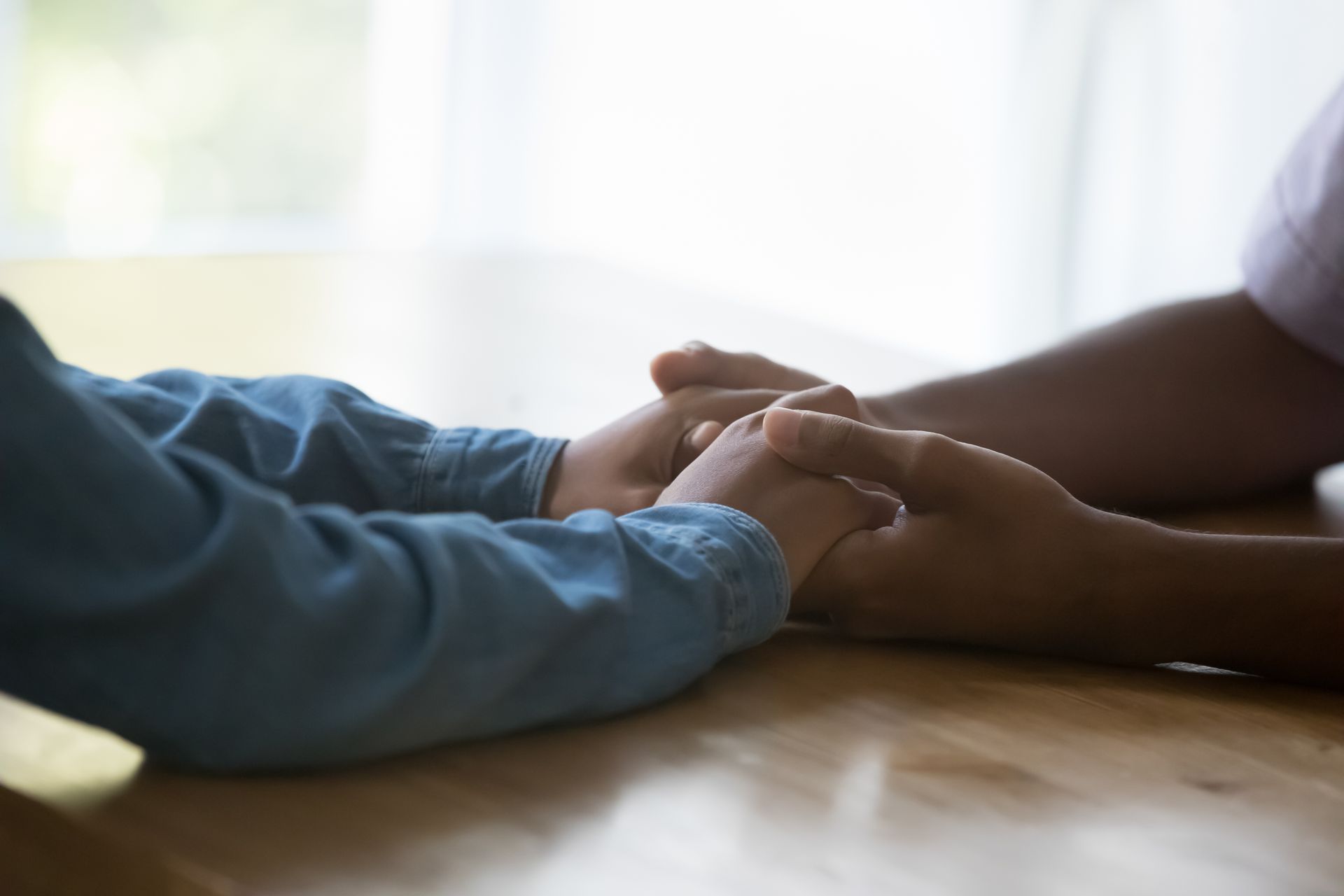 Hands gently clasped together on a wooden table near a bright window.