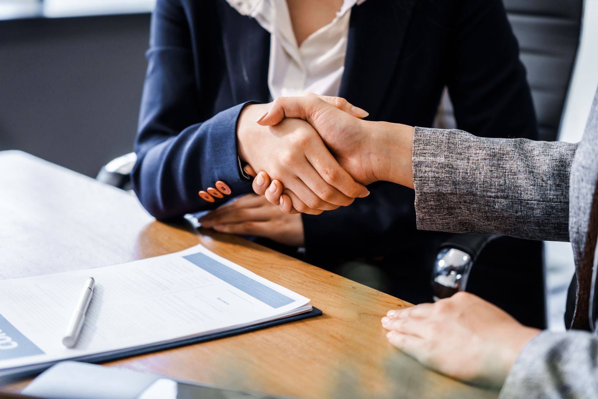 Two people shaking hands over a desk with papers and a pen, in a business meeting