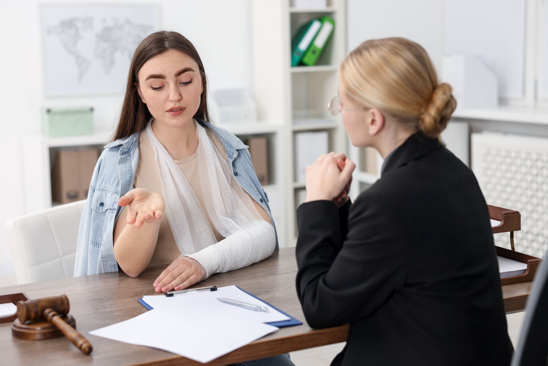 Two women in a meeting, one showing an injured arm across a desk with papers and a gavel