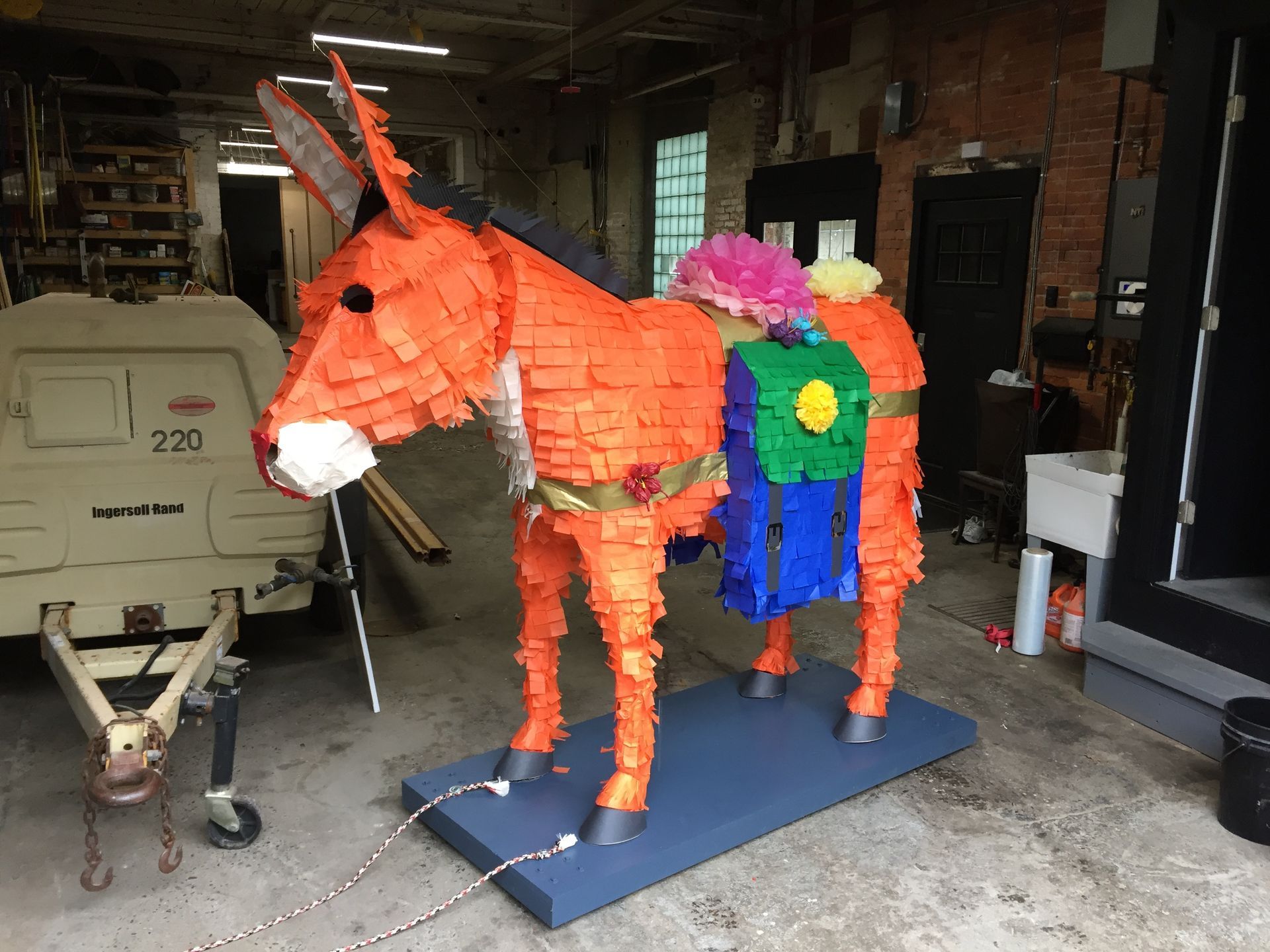 Orange donkey piñata with colorful saddle on a blue platform in a garage.