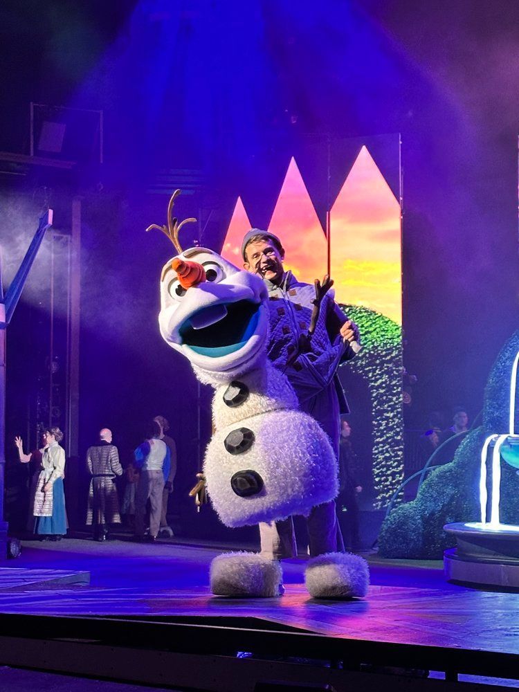 A man is singing into a microphone next to a stuffed snowman on a stage.