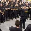 Un groupe important de personnes en uniforme noir écoute une personne qui gesticule, à l'intérieur d'un bâtiment.