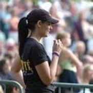 Femme en casquette et chemise noires, queue de cheval, tenant quelque chose, devant une foule.