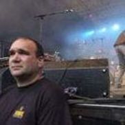 Homme en chemise noire à un concert en plein air, regardant vers la gauche, la scène et les haut-parleurs étant flous en arrière-plan.