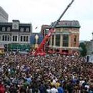 Foule importante rassemblée à l'extérieur, grue rouge en arrière-plan. Bâtiments en arrière-plan.
