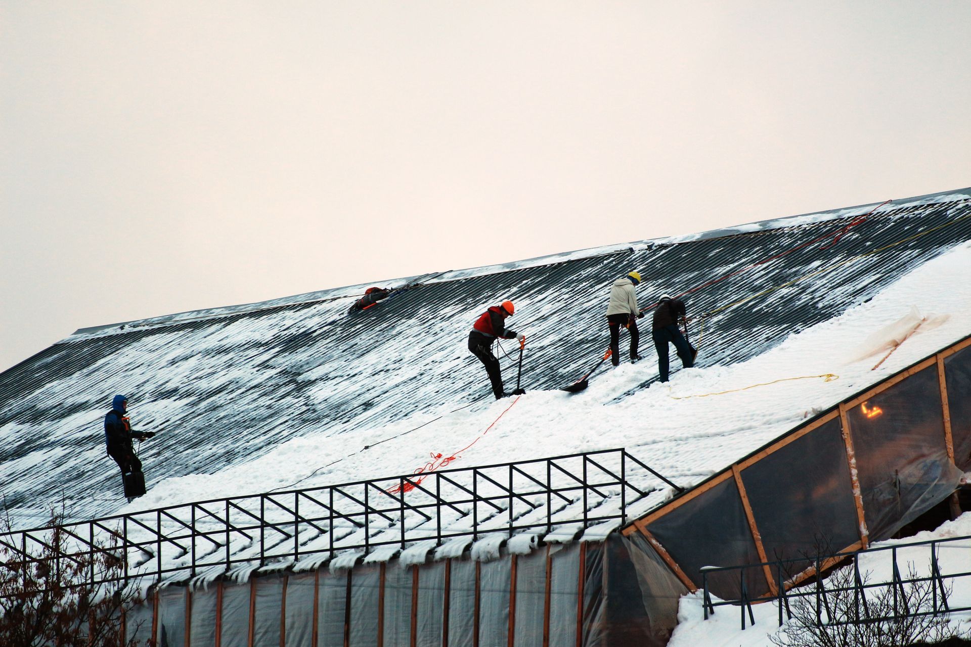 Three people climb a snowy ski slope near a fenced path on a cloudy day