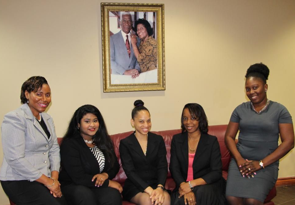 Five people pose indoors beneath a framed portrait, standing and sitting in a beige room.