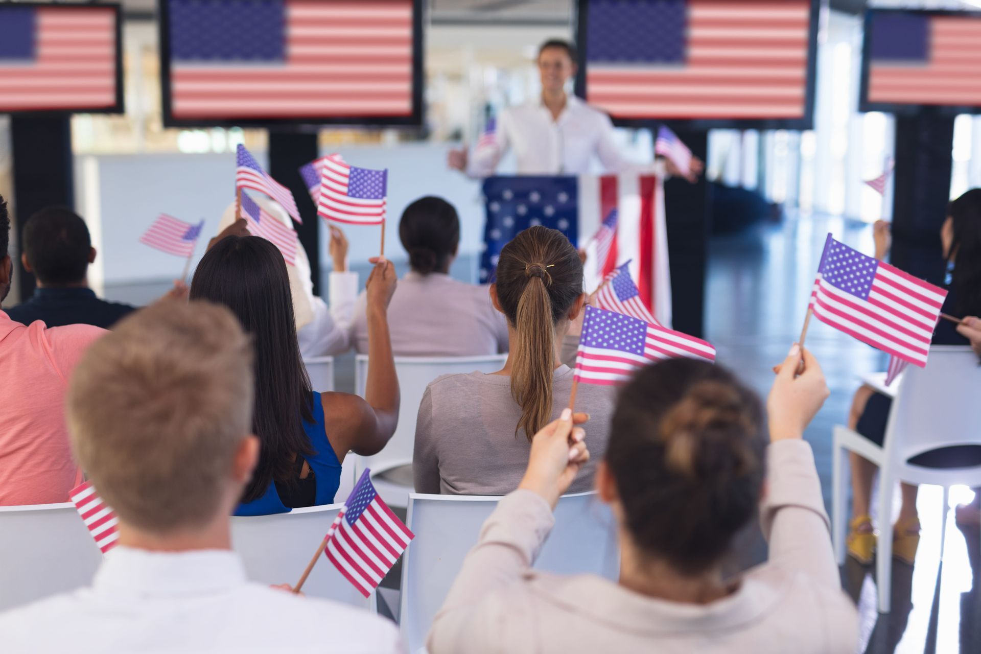Audience waving U.S. flags at a political rally with a speaker onstage and American flags in the background