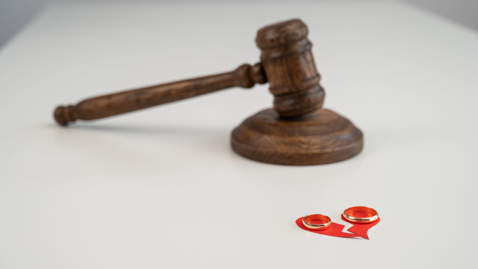 A wooden gavel sits on a white surface above two wedding rings resting on a small, torn red paper heart.