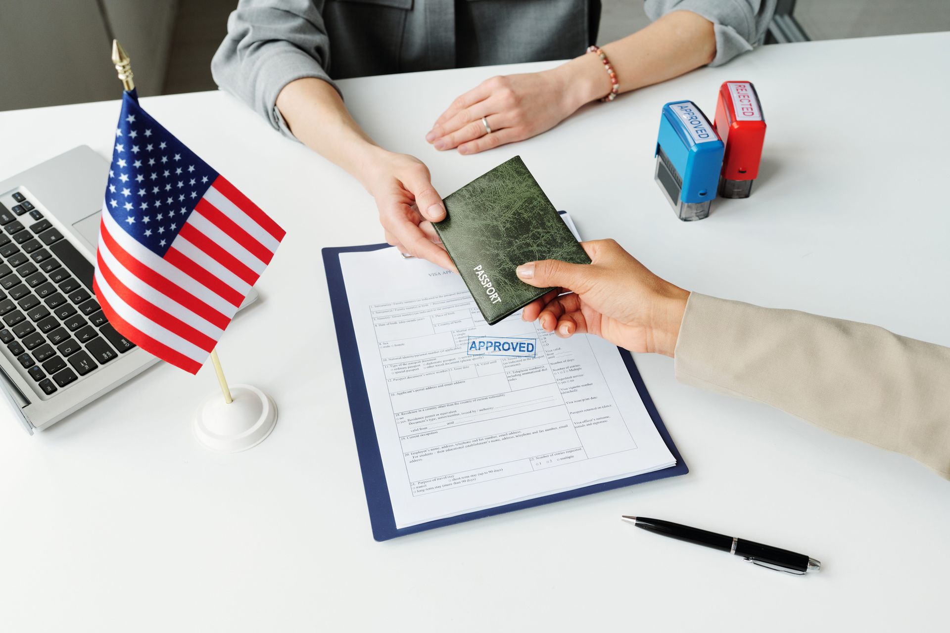 Hands exchanging a passport over a customs form with a small U.S. flag and passport stampers on a desk