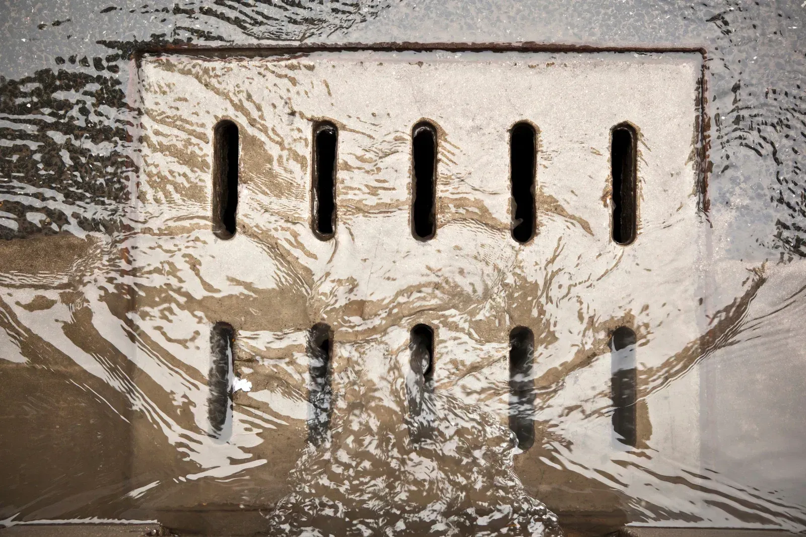 Water flowing over a metal grate with vertical slots, splashing on a gray surface.