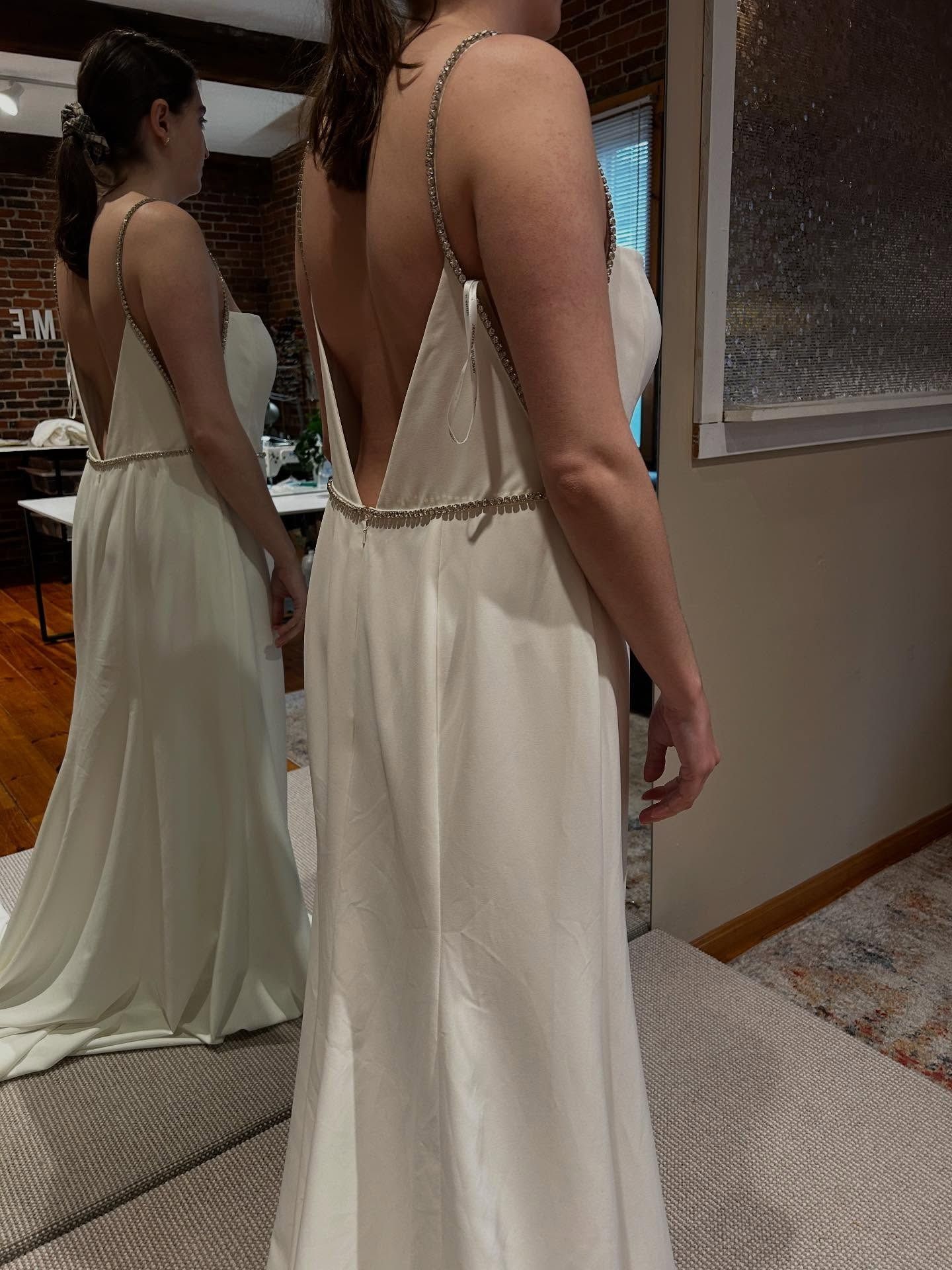 Woman in a backless white dress standing by a mirror in a room, showing the dress’s draped train.