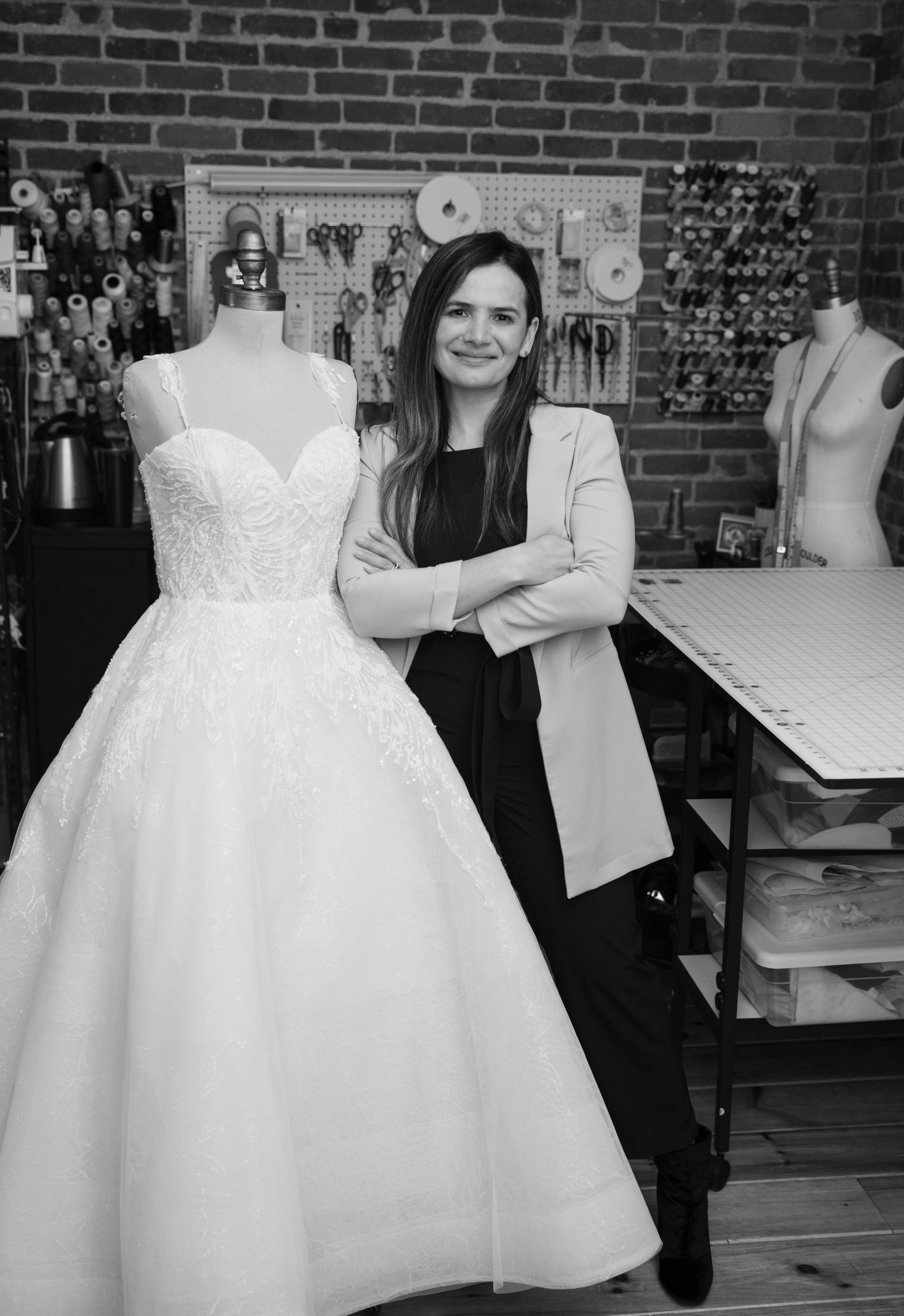 Woman standing beside a strapless white wedding gown in a dress shop, smiling with arms crossed.