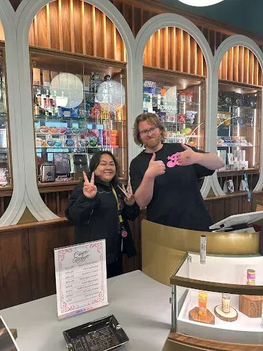 Two people posing behind a display counter in a shop, smiling and holding small pink items.