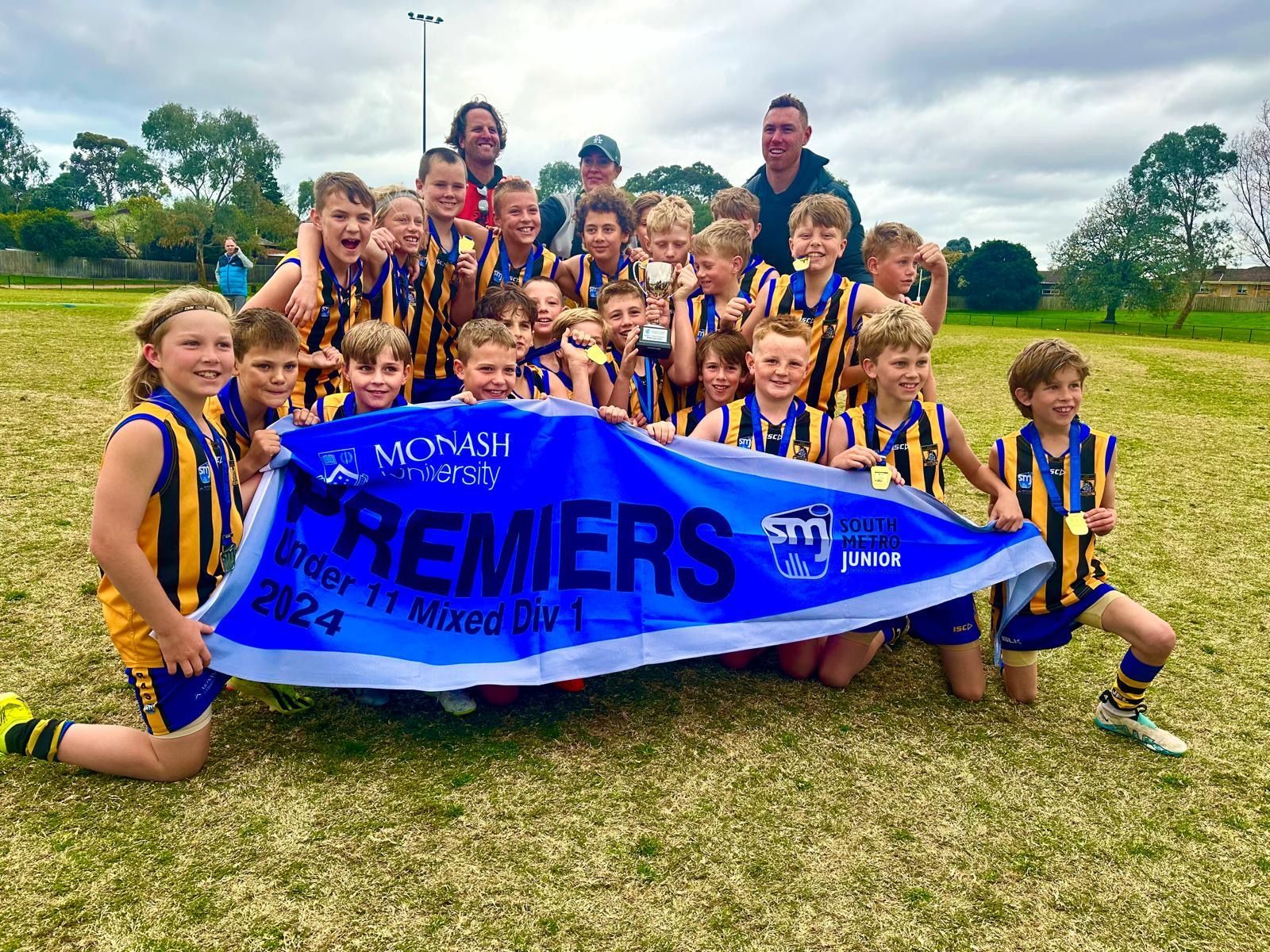 A youth Australian rules football team in gold and navy uniforms celebrates holding a 2024 premier banner on a grassy field.