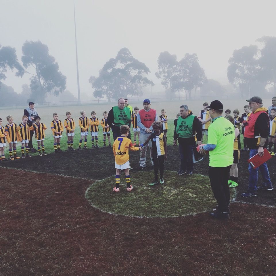 Two young players shake hands in a center circle on a foggy sports field, surrounded by teammates, coaches, and officials.