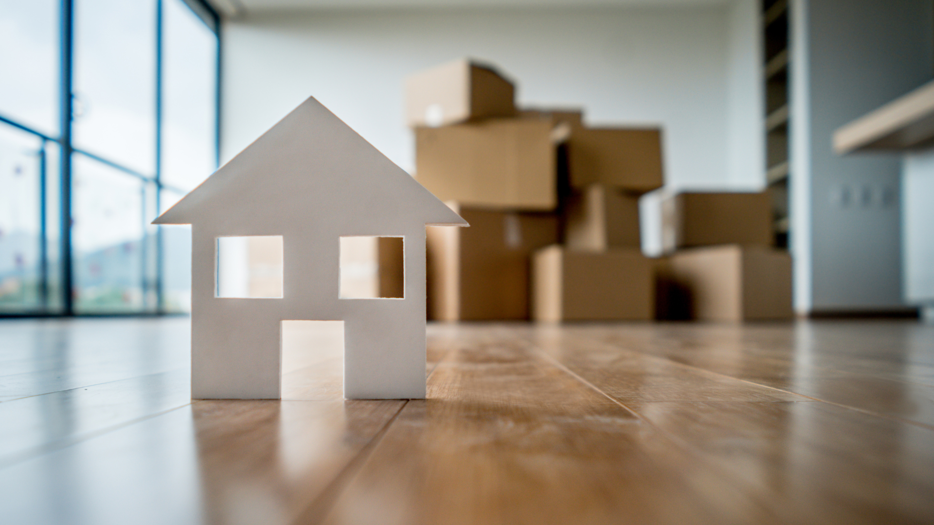 Cardboard house cutout on hardwood floor; moving boxes in the background.