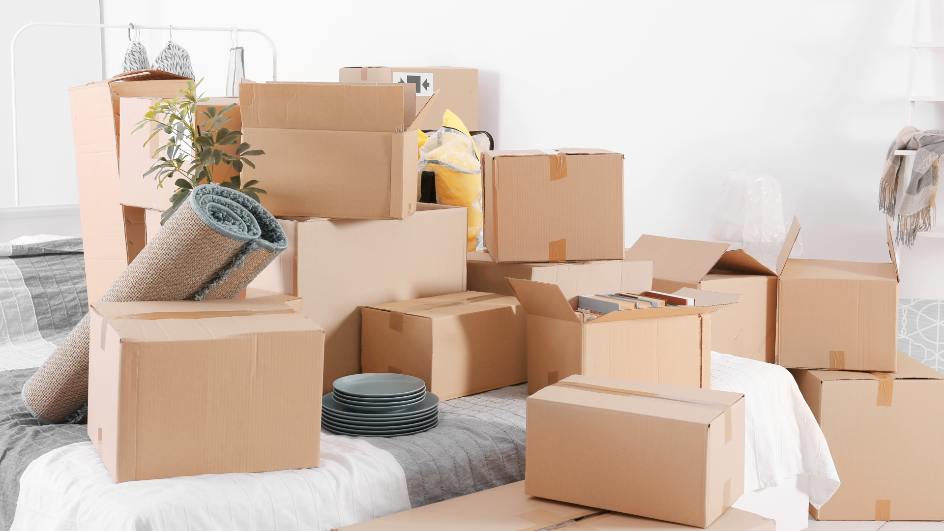 Pile of cardboard boxes on a bed, suggesting a home move. A rolled rug and a robot vacuum are visible too.