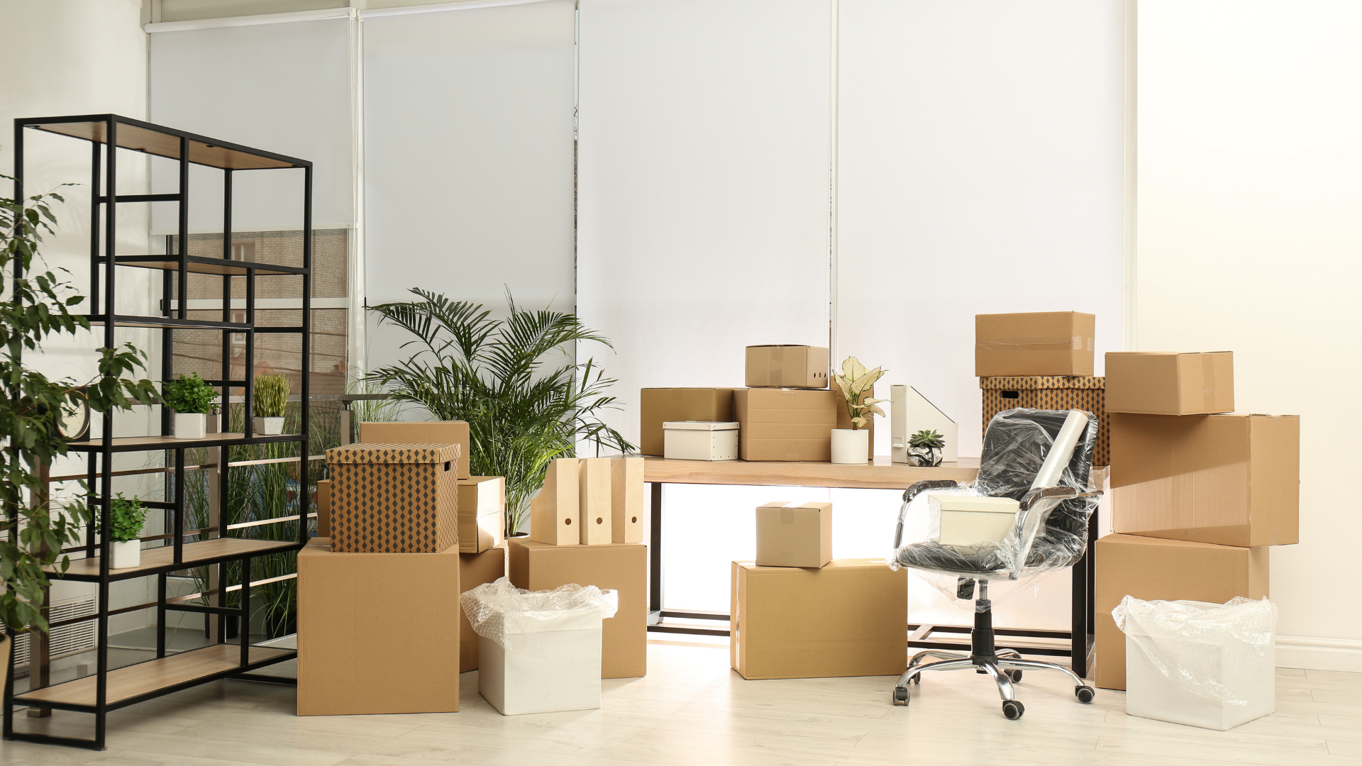 Office space packed for moving, cardboard boxes stacked, desk, shelving, chair.