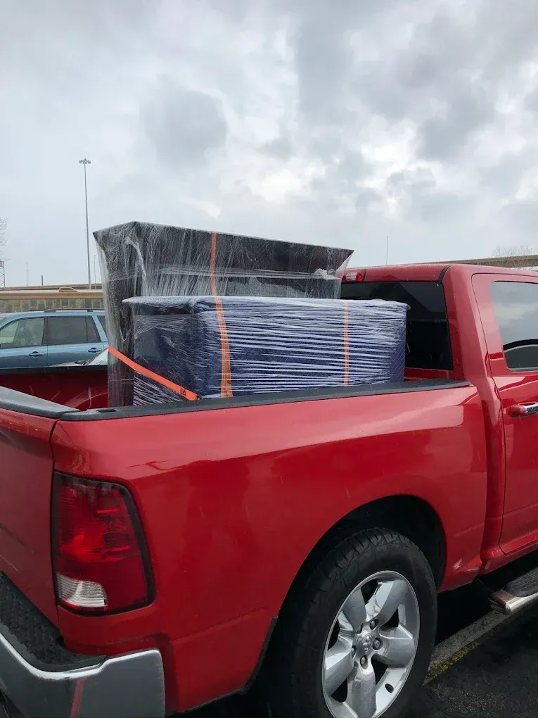 Red pickup truck bed loaded with wrapped furniture, secured by orange straps. Overcast sky.