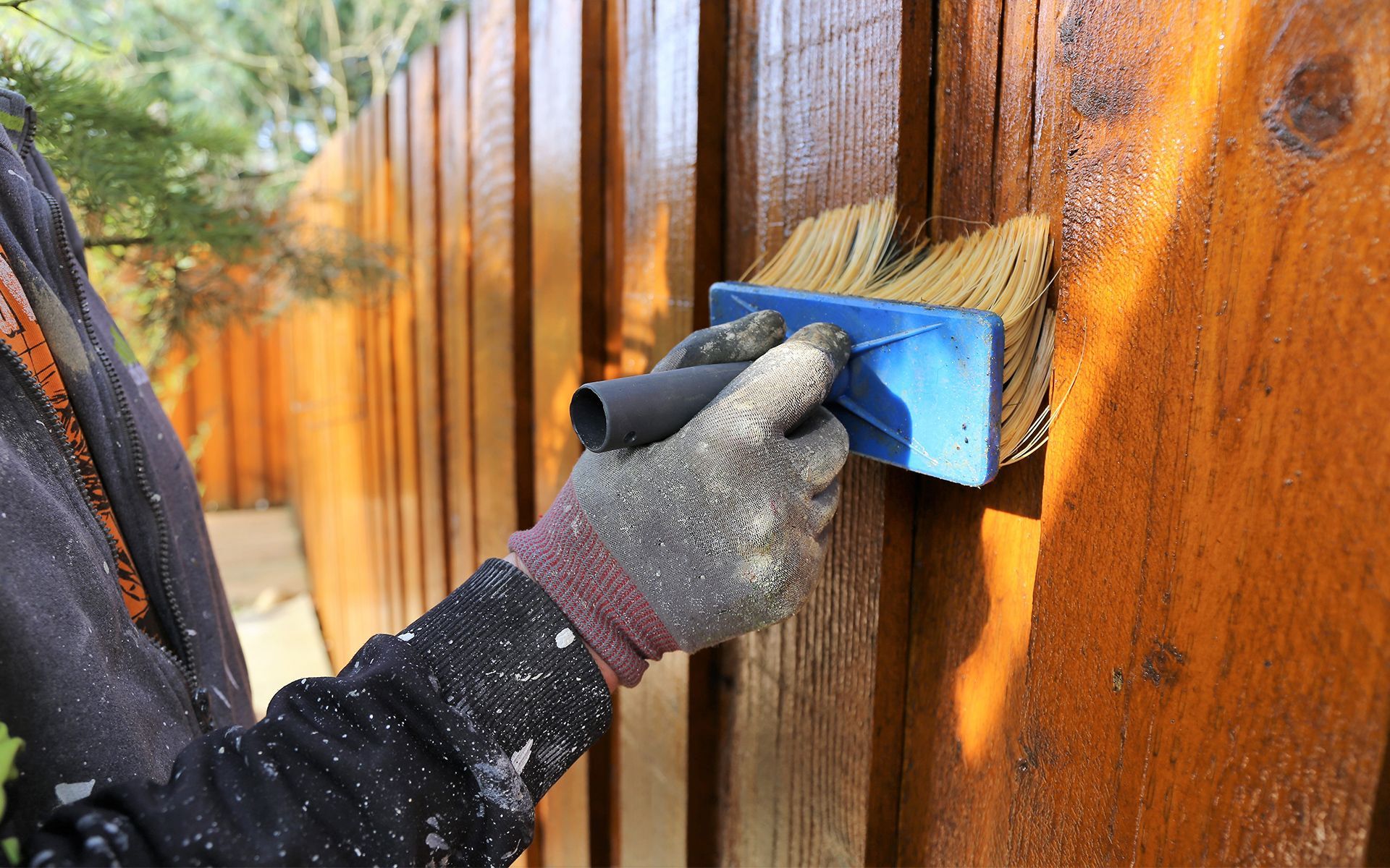 Person staining a wooden fence with a brush, wearing gloves, in outdoor setting.