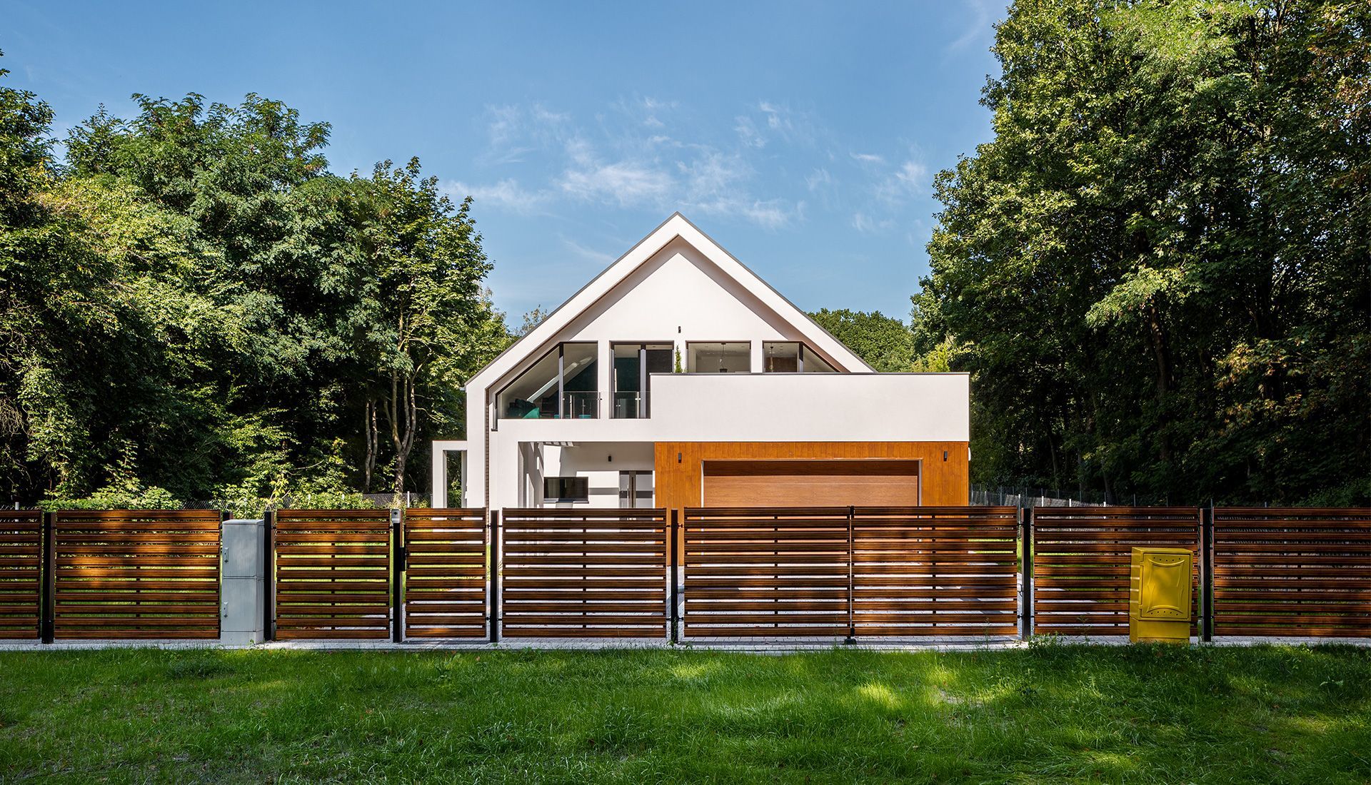 Modern white house with a gabled roof behind a wooden fence and trees.