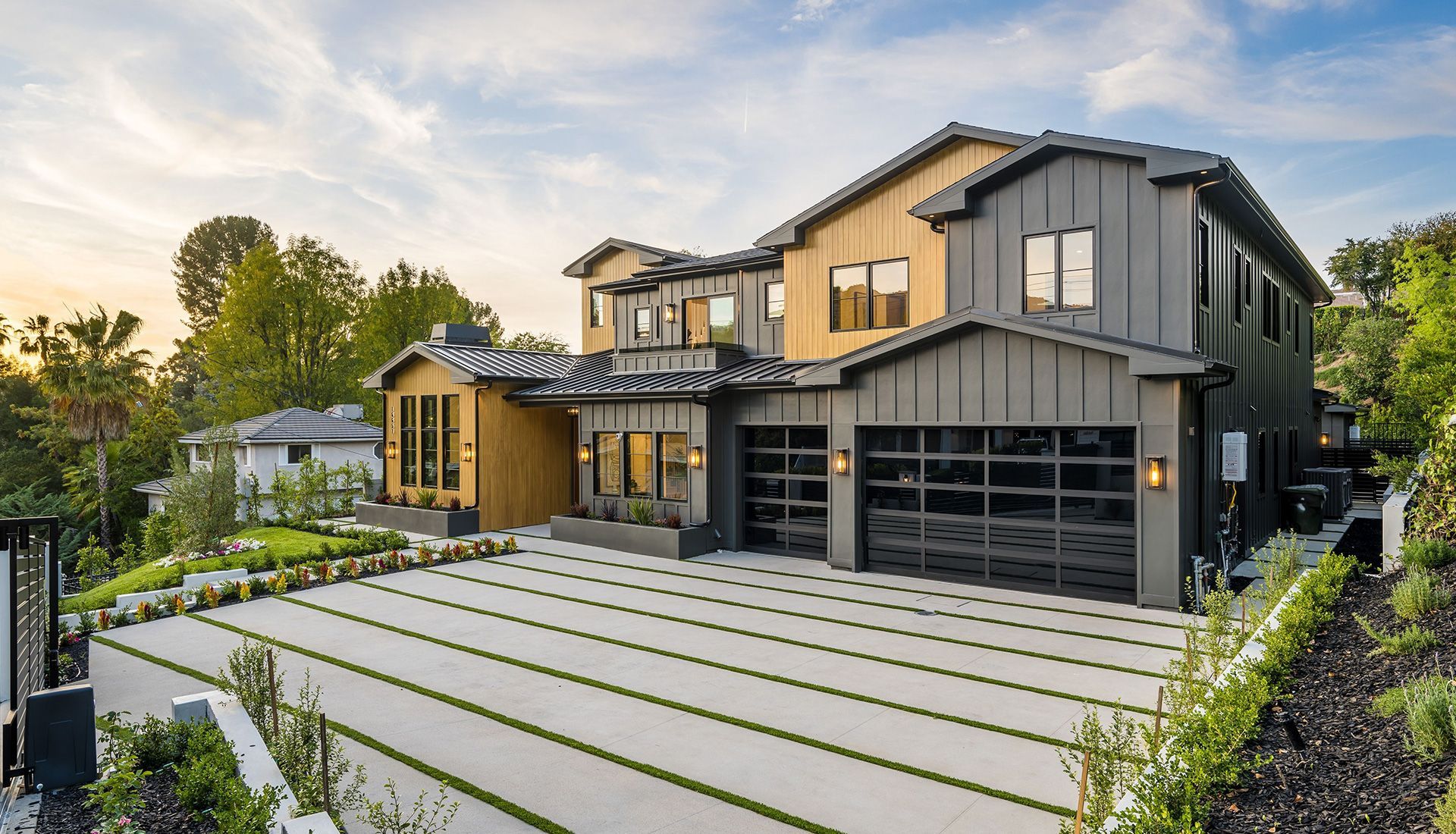 Modern two-story house with gray and wood siding, glass garage doors, and a long driveway with grass strips.