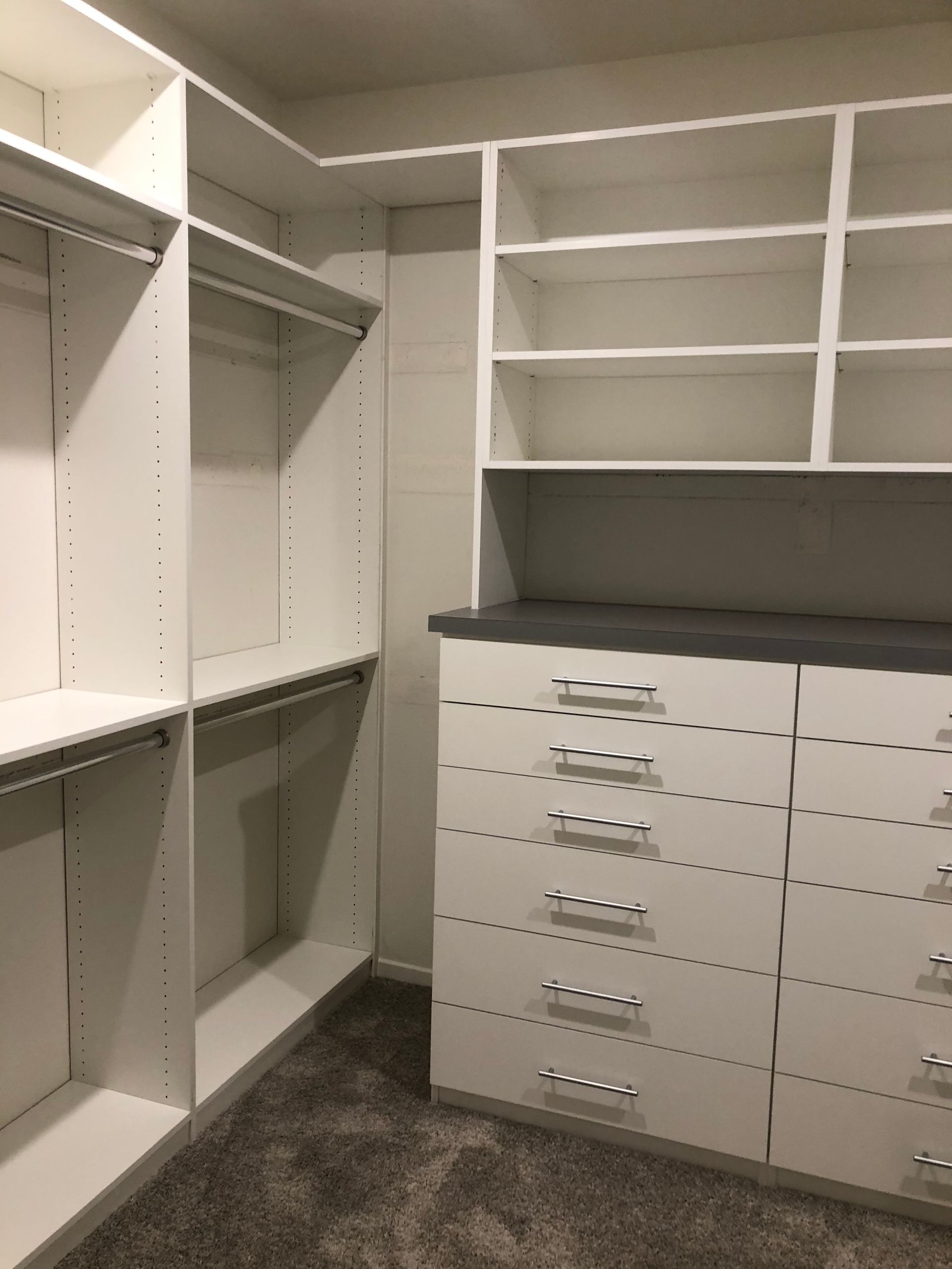 Empty white walk-in closet with shelves, drawers, and hanging rods on gray carpet.