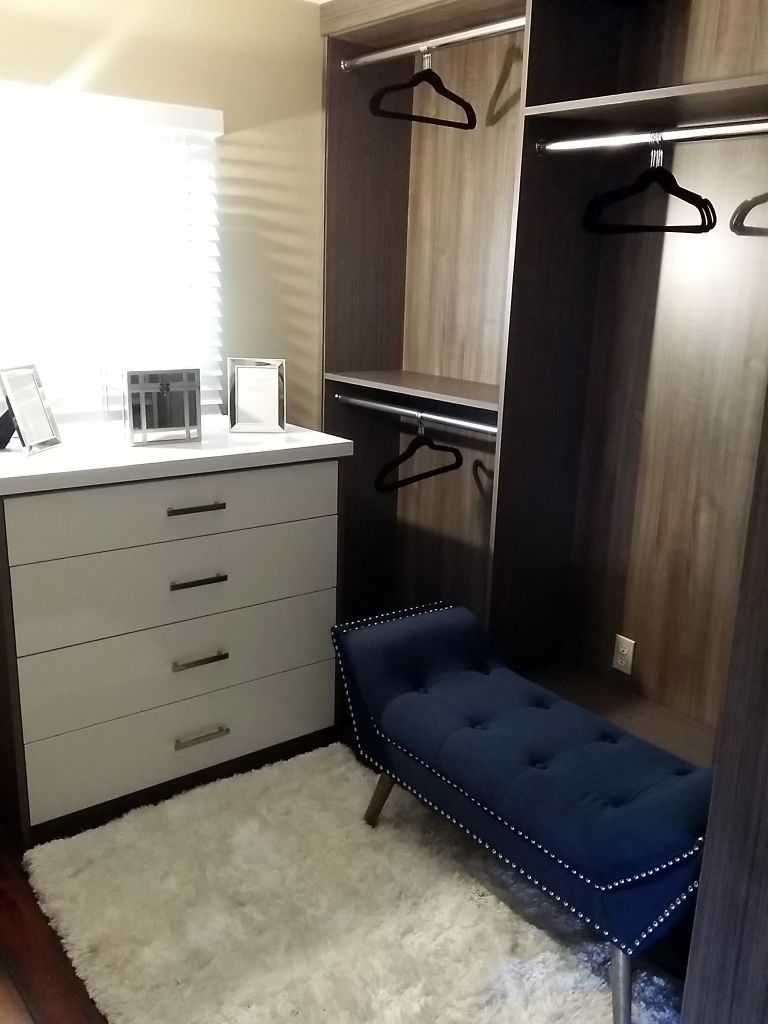 Open closet with drawers, hanging rods, and blue upholstered bench beside a white dresser near a window.