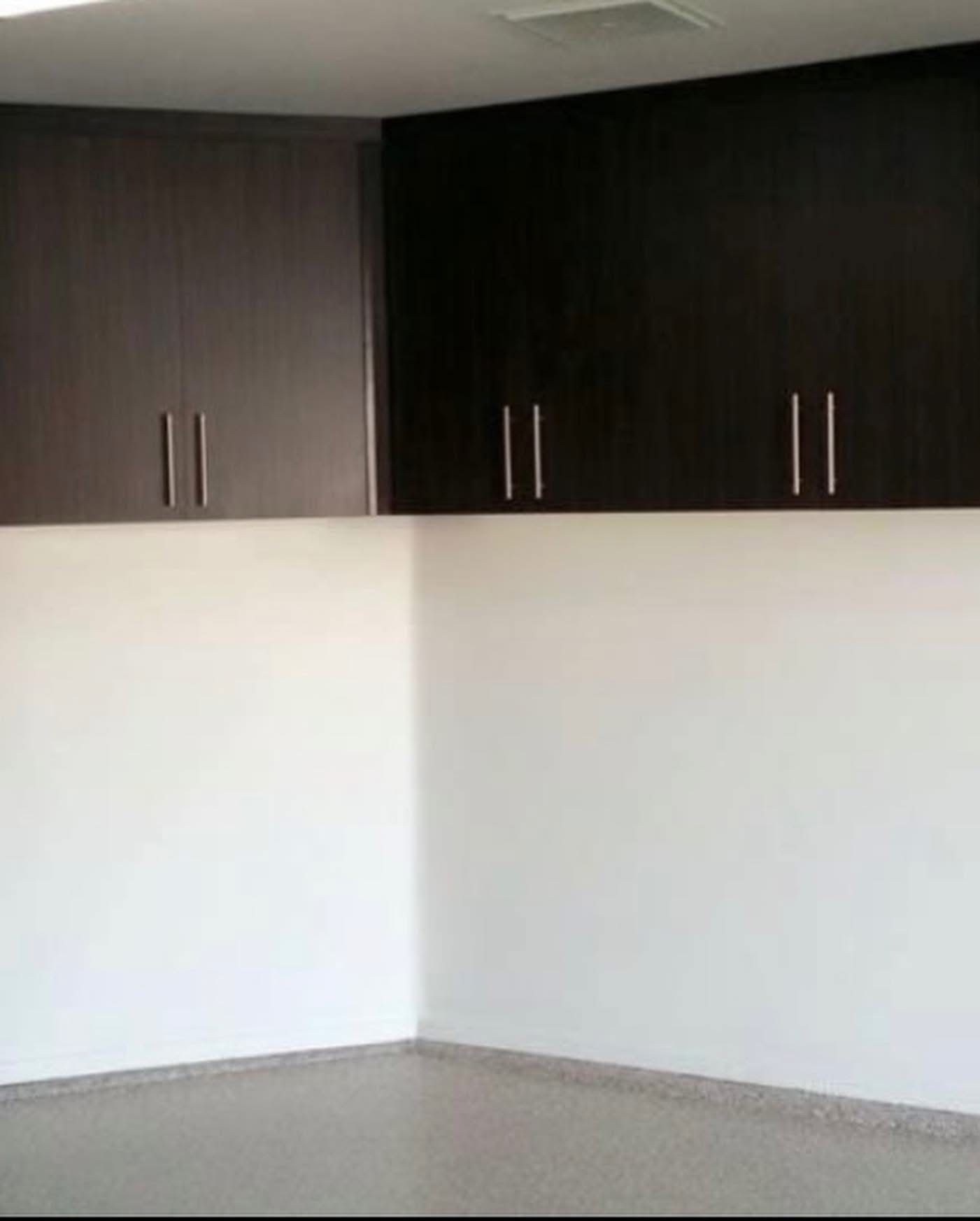 Empty room corner with white walls, gray floor, and dark wood cabinets mounted high on the wall.