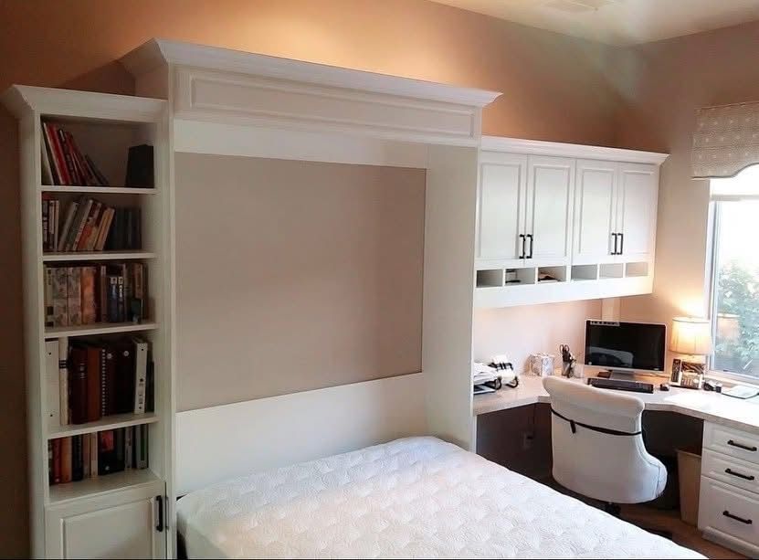 Built-in desk nook with white cabinetry, bookshelf, chair, and bed in a small bedroom/office nook